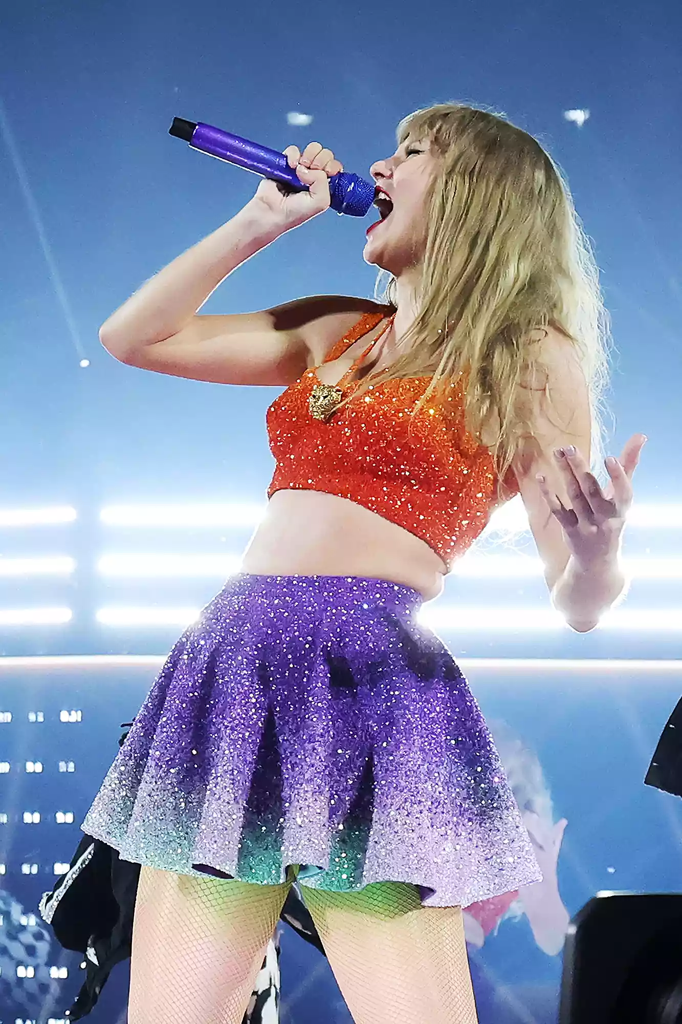 Taylor Swift singing on stage during the 'Eras Tour,' dressed in an orange sequined crop top and a purple glittery skirt. The eye-catching outfit embodies the energetic and dynamic aesthetic of her '1989' album era, complete with a matching purple microphone.