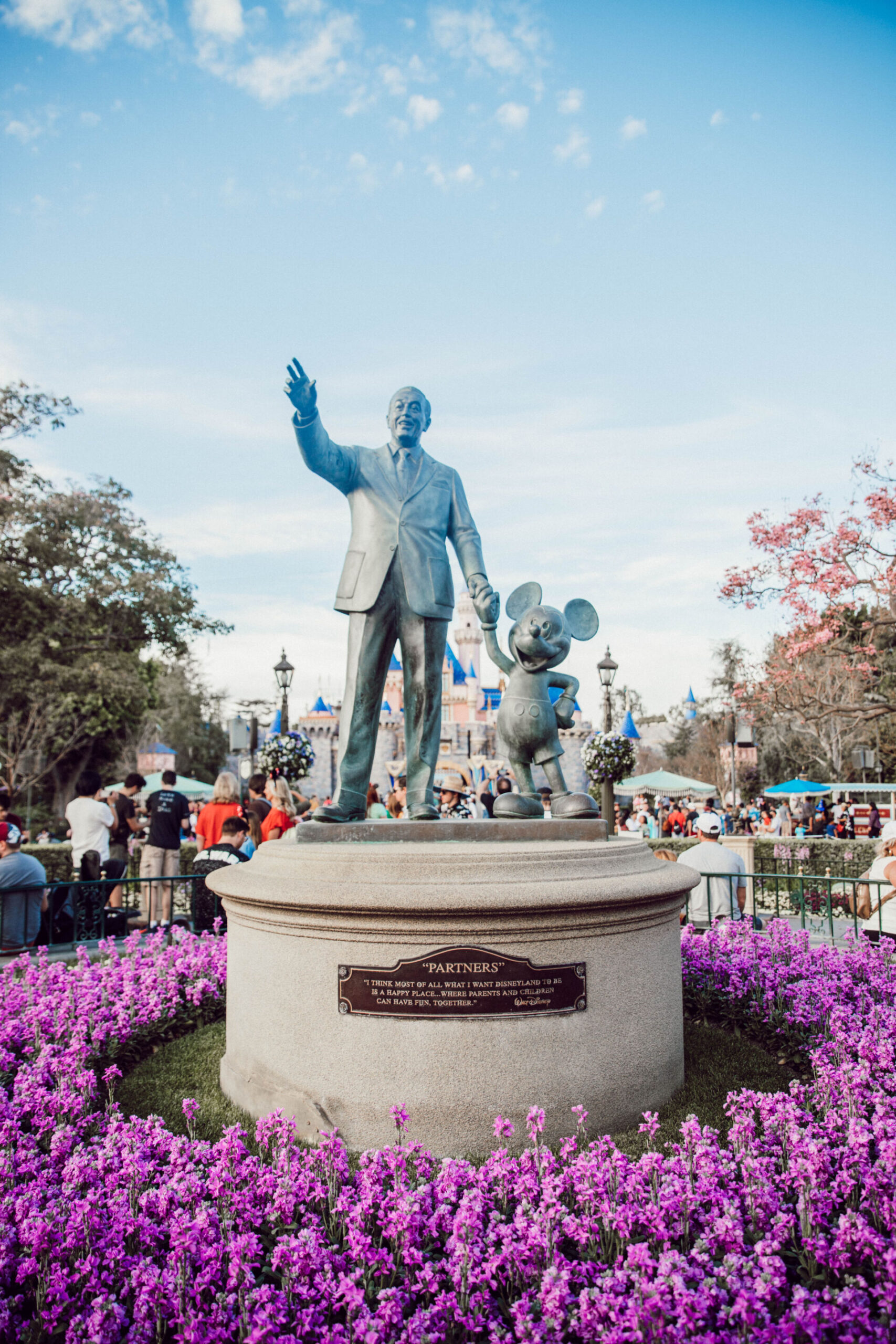 Partners statue of Walt Disney holding hands with Mickey Mouse in front of Sleeping Beauty Castle at Disneyland California, surrounded by purple flowers and visitors.