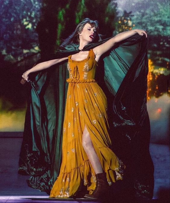 Taylor Swift performing on stage during the 'Eras Tour,' wearing a mustard yellow dress with floral embroidery and a green cloak, reflecting the enchanted and woodsy essence of her 'Evermore' album era.