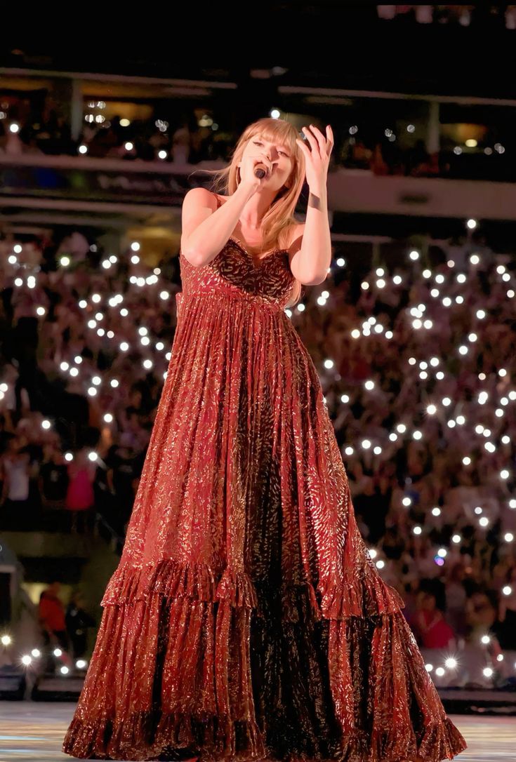 Taylor Swift singing on stage during the 'Eras Tour,' dressed in a shimmering red and gold gown, capturing the warm and autumnal style of her 'Evermore' album era.