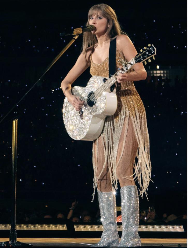 Taylor Swift performing on stage during the 'Eras Tour,' dressed in a shimmering gold fringe dress paired with sparkling silver knee-high boots. She plays a glittery silver guitar while singing into a golden microphone, capturing the glamorous and nostalgic essence of her 'Fearless' album era, with a backdrop of twinkling stage lights.