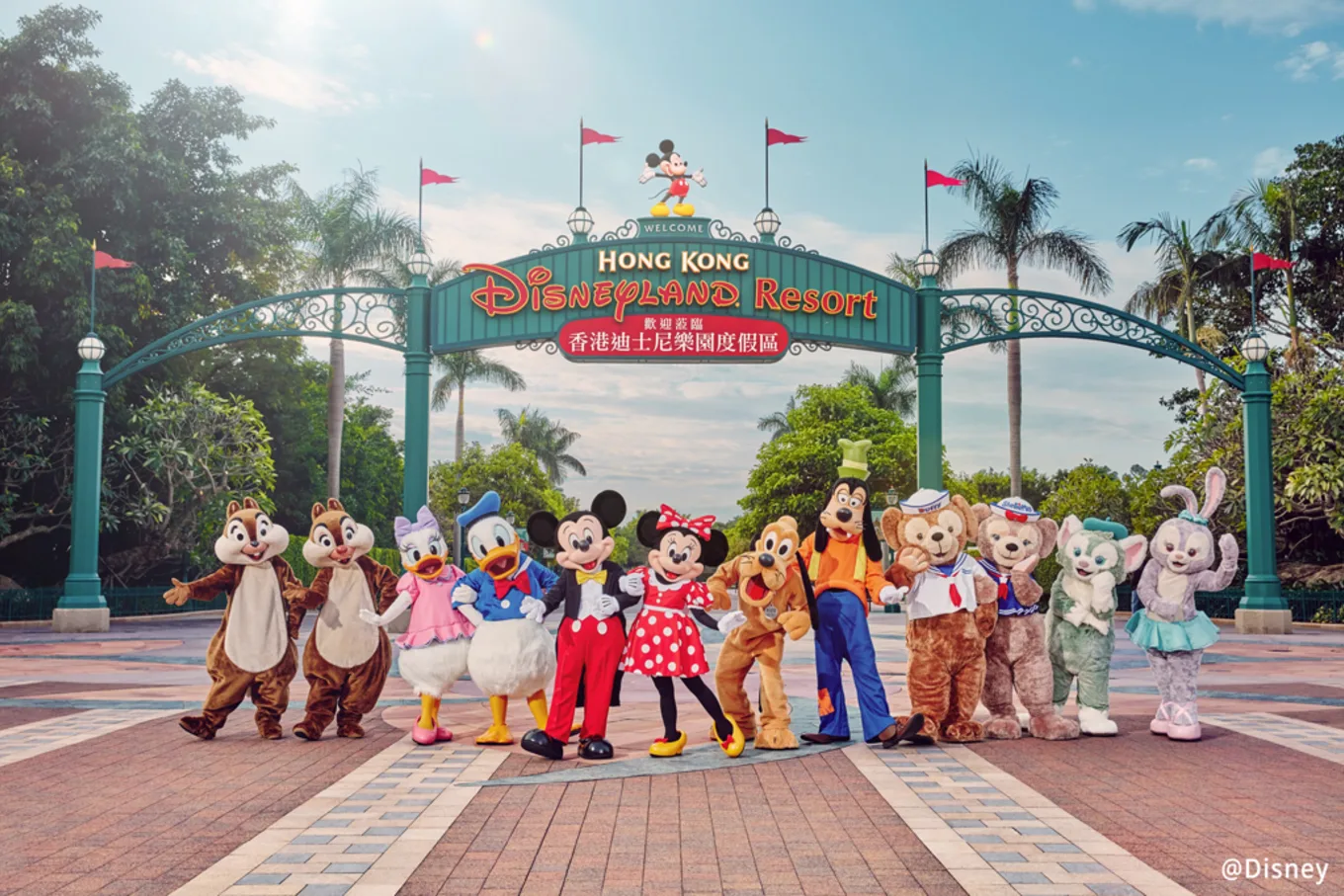 A group photo of iconic Disney characters including Mickey Mouse, Minnie Mouse, Donald Duck, Daisy Duck, Goofy, Pluto, and others standing in front of the entrance gate to Hong Kong Disneyland Resort, with vibrant greenery and clear blue skies in the background.