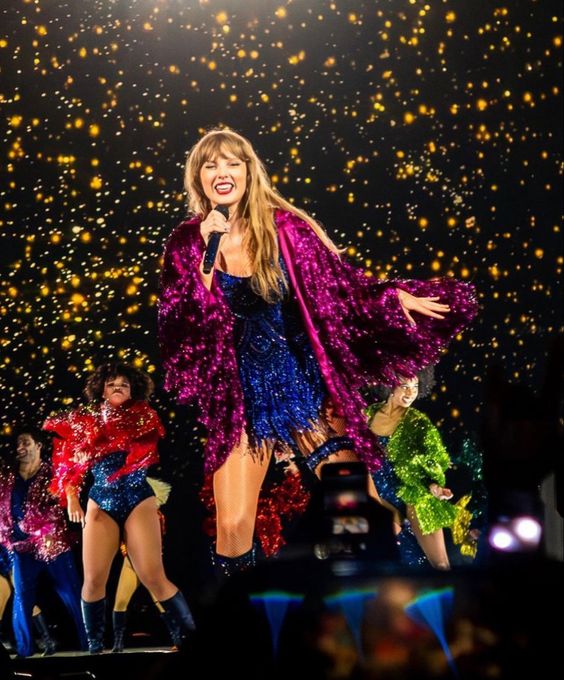 Taylor Swift performing on stage during the 'Eras Tour,' wearing a sparkly navy blue romper with a magenta cape, capturing the vibrant and colorful style of her 'Midnights' album era.