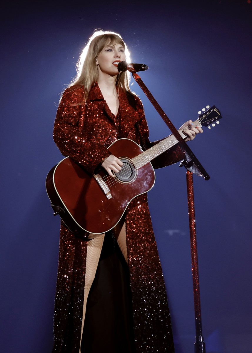 Taylor Swift performing on stage during the 'Eras Tour,' wearing a stunning red sequined long coat and playing a matching red acoustic guitar. The outfit, paired with a glittery red microphone, captures the dramatic and bold essence of her 'Red' album era.