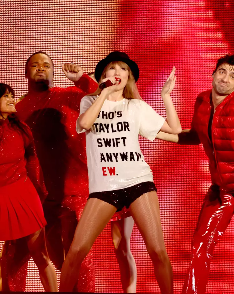 Taylor Swift on stage during the 'Eras Tour,' wearing a white t-shirt with the phrase 'Who's Taylor Swift Anyway? EW.' along with black shorts and a black hat. She performs energetically alongside her dancers dressed in red, capturing the playful and iconic moments from her 'Red' album era.