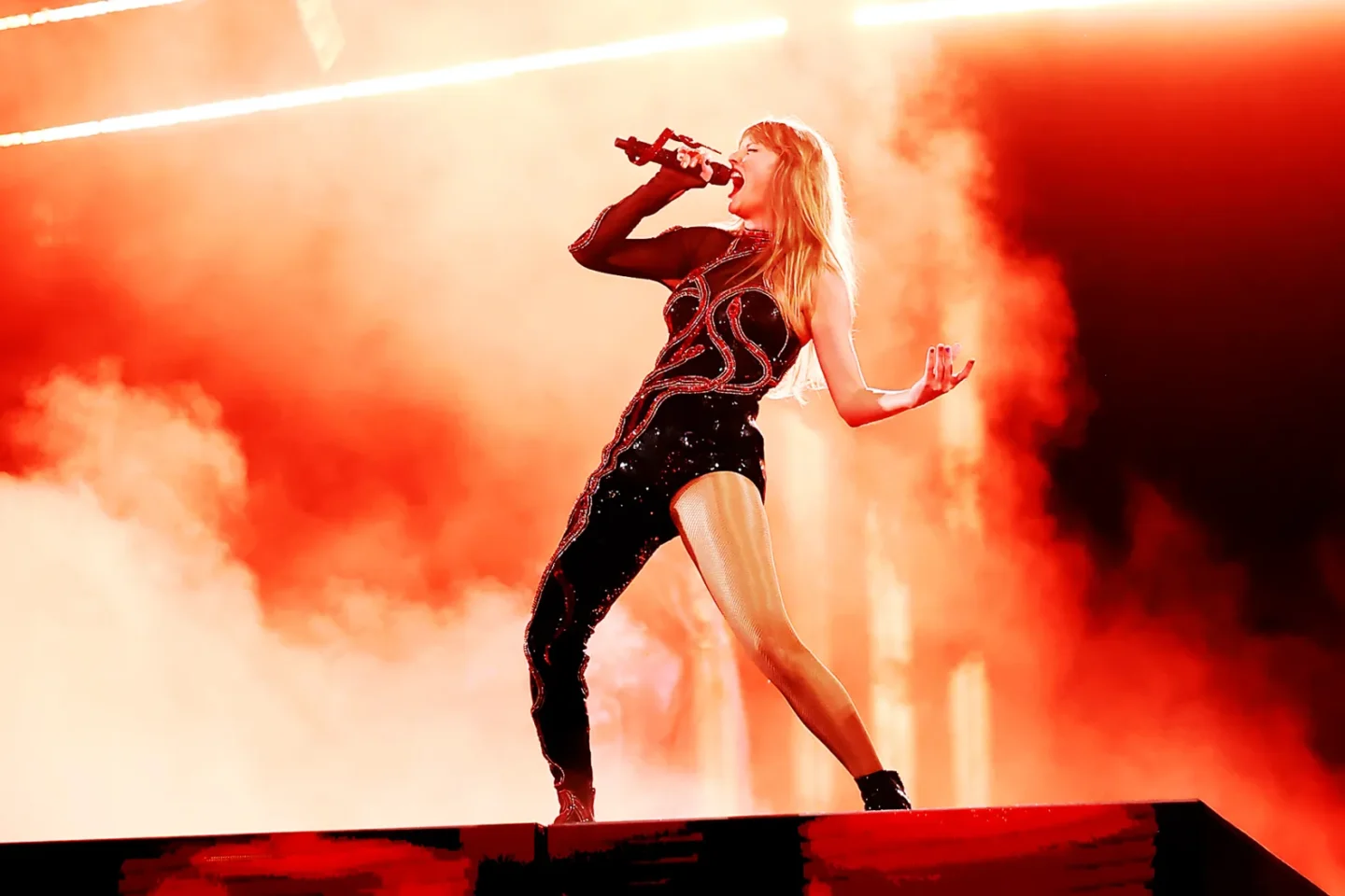 Taylor Swift performing on stage during the 'Eras Tour,' wearing a black sequined bodysuit with red snake-like patterns and a sheer sleeve. The dramatic outfit is highlighted by a fiery red and orange background, capturing the bold and edgy essence of her 'Reputation' album era.