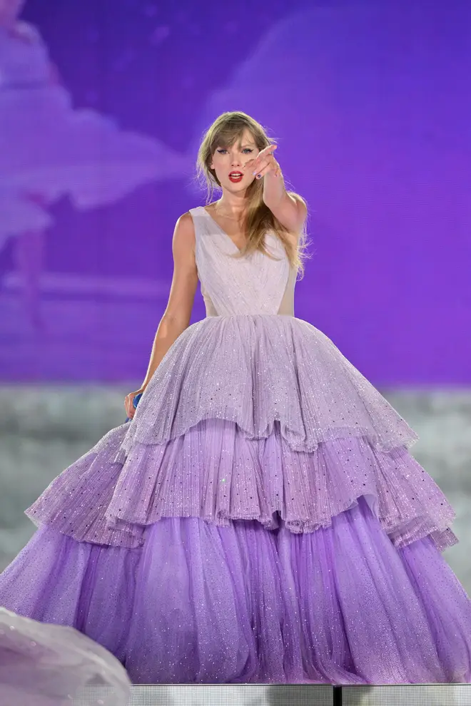 Taylor Swift performing on stage during the 'Eras Tour,' wearing a stunning lavender ball gown with multiple tiered layers and sparkly embellishments. The dress features a fitted bodice and a voluminous skirt, capturing the whimsical and fairytale-like essence of her 'Speak Now' album era, set against a vibrant purple backdrop.