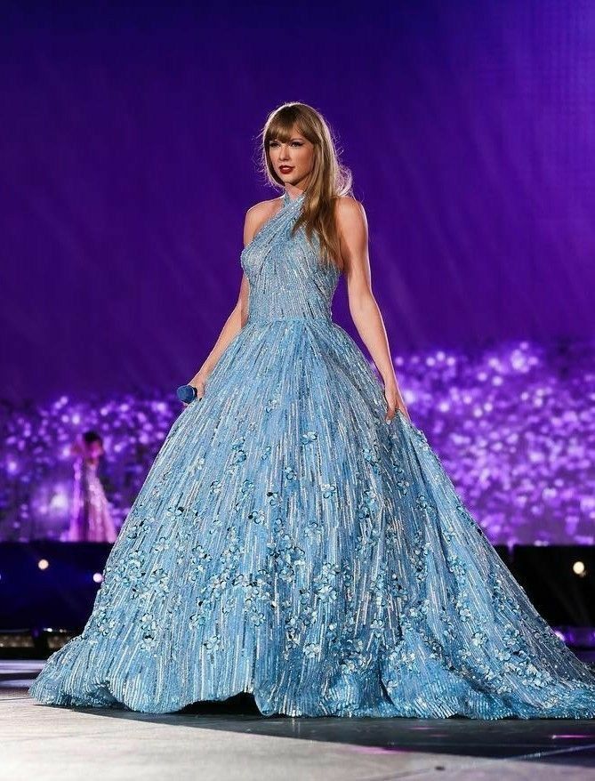 Taylor Swift performing on stage during the 'Eras Tour,' wearing a breathtaking blue ball gown with a halter neckline, intricate floral appliqués, and shimmering fabric. The dress captures the ethereal and fairytale-like essence of her 'Speak Now' album era, set against a vivid purple backdrop that enhances the magical ambiance of her performance.